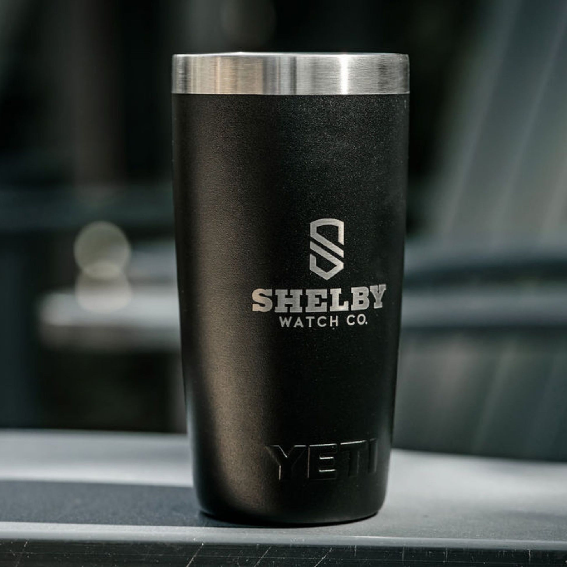 A matte black YETI travel tumbler with a brushed stainless steel rim, featuring the Shelby Watch Co. logo printed in silver on the front, set against a blurred dark background.
