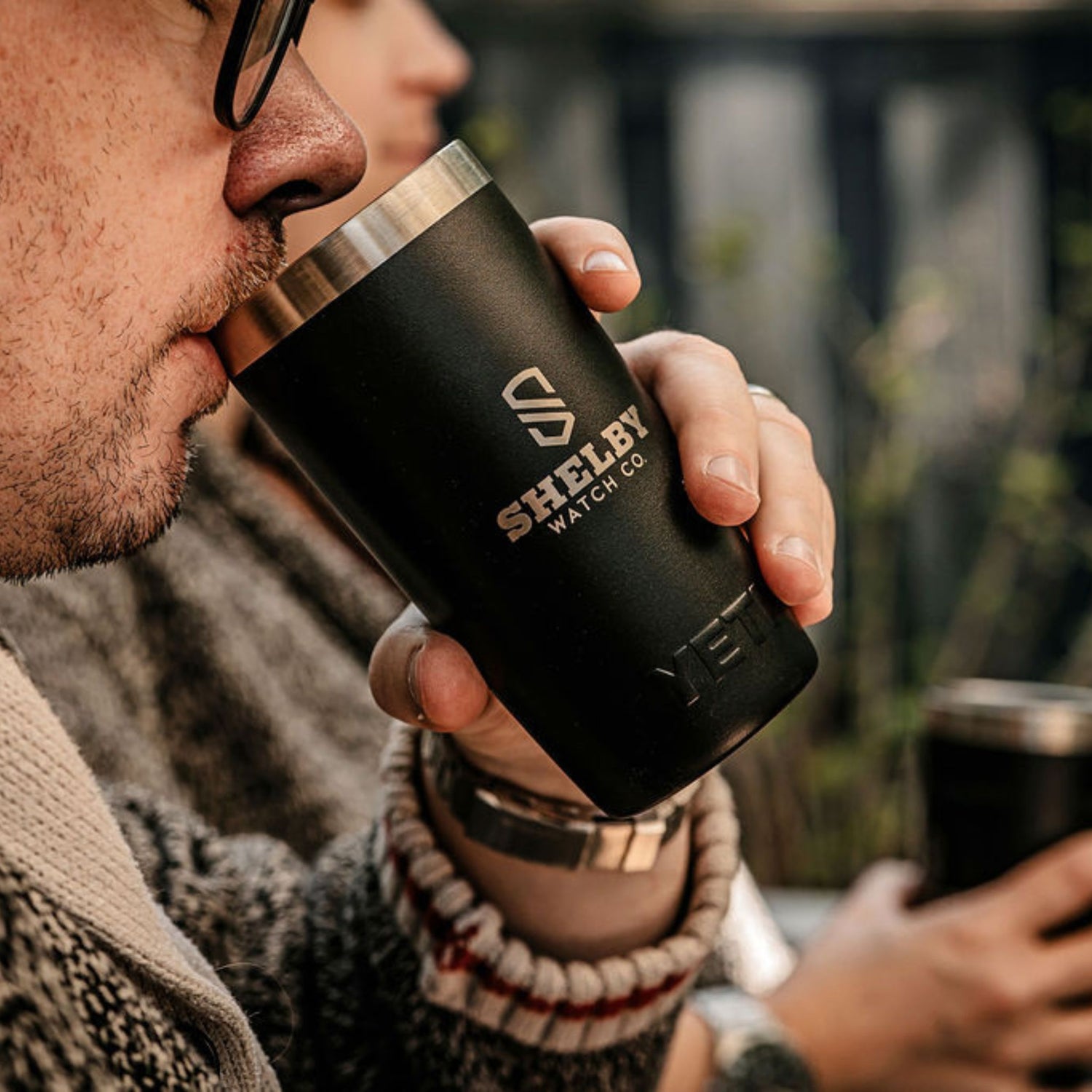A close-up lifestyle shot of a man drinking from a matte black insulated travel tumbler with a stainless steel rim and Shelby Watch Co. engraved in silver. 