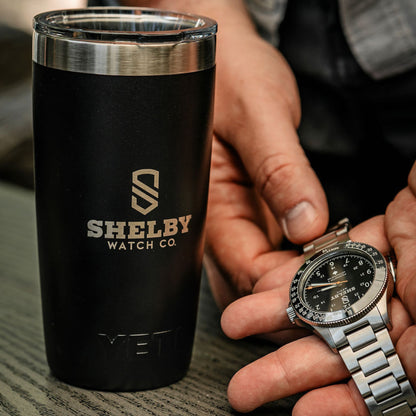 A black stainless steel tumbler with a silver rim Shelby Watch Co. branding, shown next to a person's hands holding a silver stainless steel watch with a black dial.