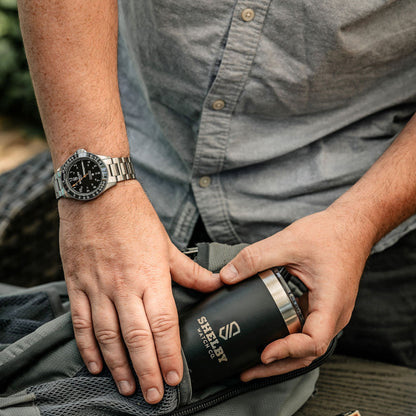 A close-up lifestyle shot of a person wearing a stainless steel watch putting a black insulated travel mug engraved with Shelby Watch Co. into a backpack. 