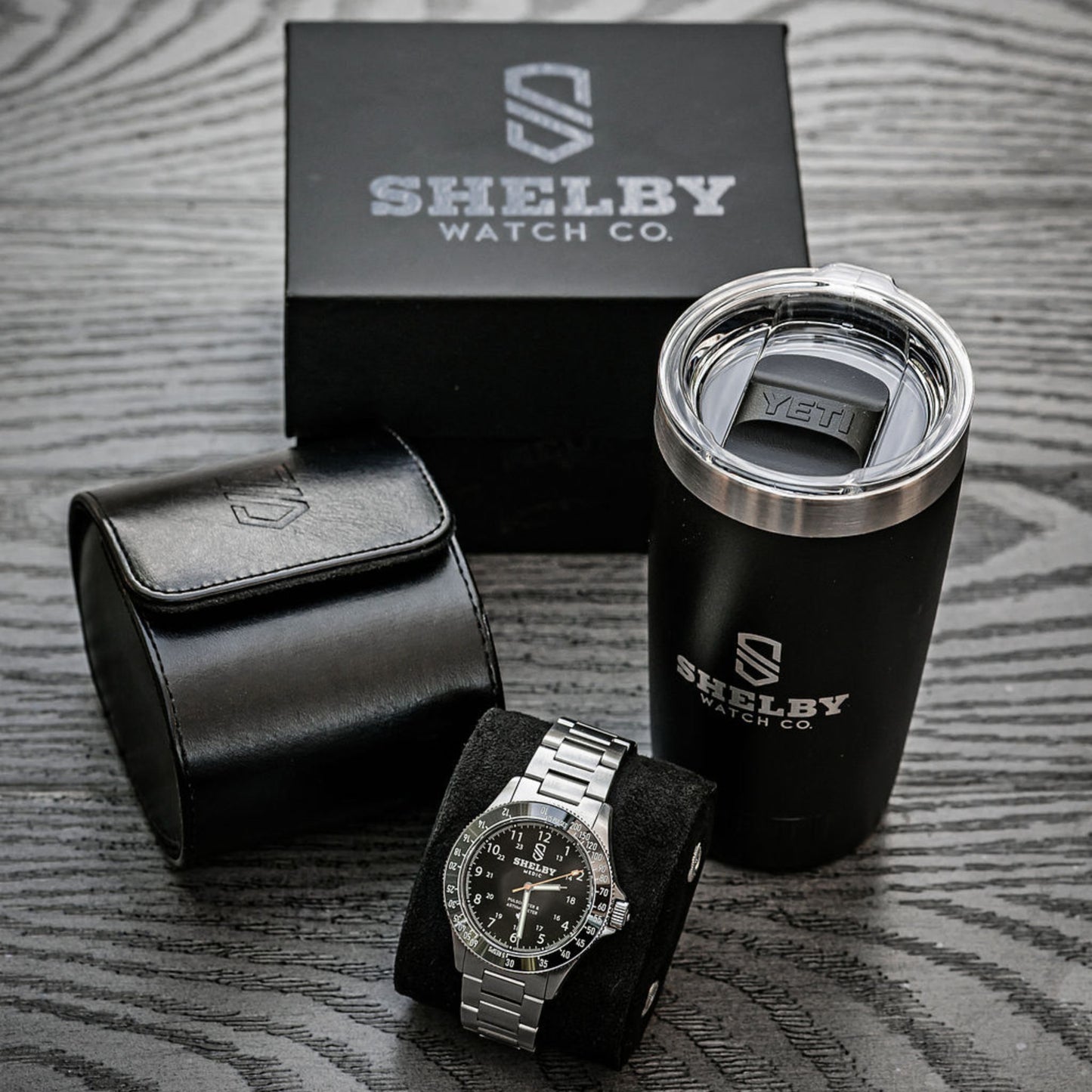 Shelby Watch Co. branded watch presentation set on a gray wood-textured surface, including a silver-tone watch on a black cushion, a black leather watch pouch, a black travel tumbler with stainless steel rim, and the black branded box behind.