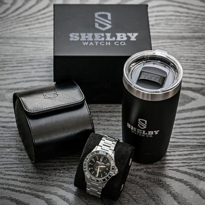 Shelby Watch Co. branded watch presentation set on a gray wood-textured surface, including a silver-tone watch on a black cushion, a black leather watch pouch, a black travel tumbler with stainless steel rim, and the black branded box behind.