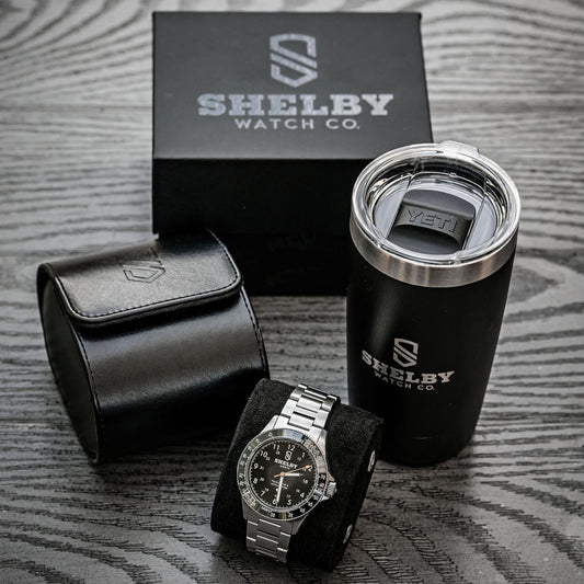 Shelby Watch Co. branded watch presentation set on a gray wood-textured surface, including a silver-tone watch on a black cushion, a black leather watch pouch, a black travel tumbler with stainless steel rim, and the black branded box behind.